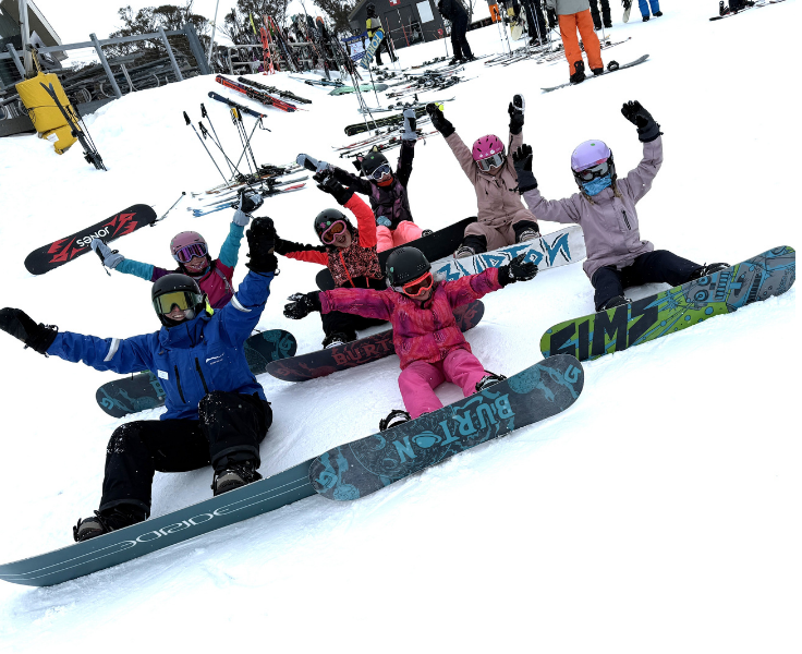 A group of students with their snowboarding instructor waving at the camera.